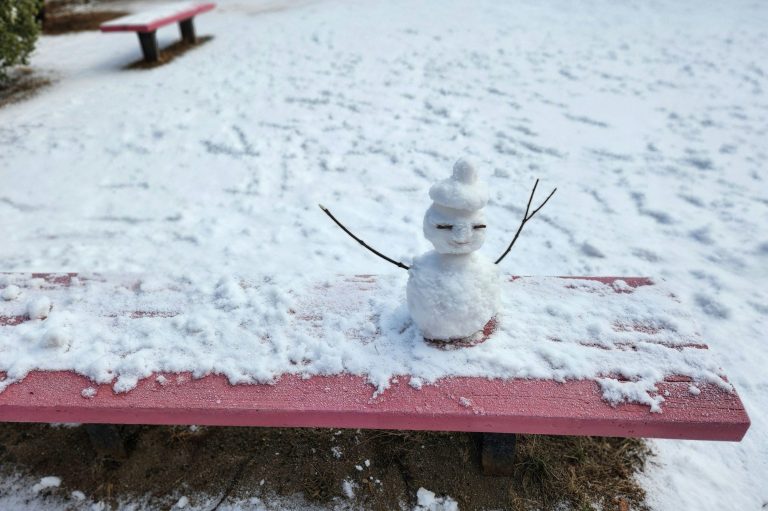 Ein kleiner Schneemann sitzt auf einer rosa Bank im Schnee.